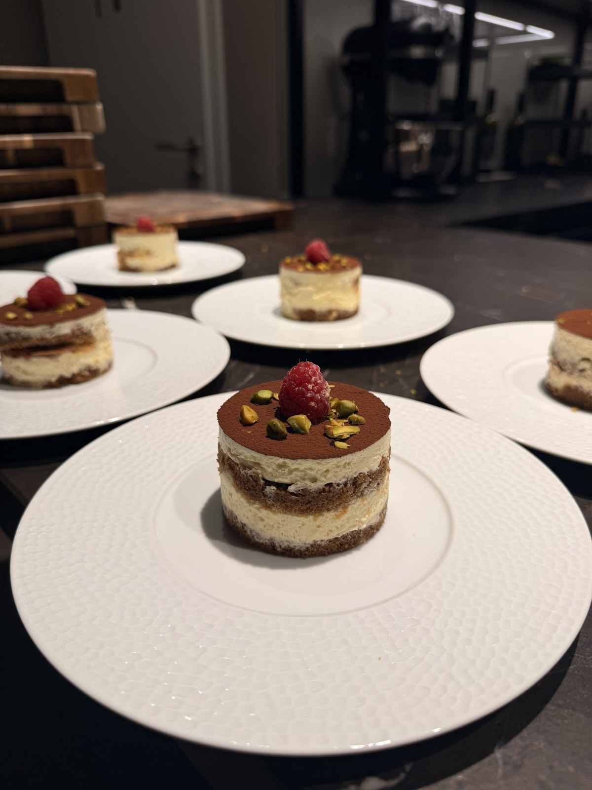 Mehrere Tiramisu-Dessertchen mit Himbeere und Pistazie auf weissen Tellern im Kochstudio